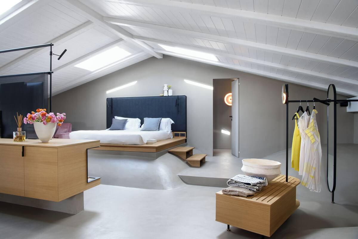 A modern loft bedroom with a skylight, a bed with a blue headboard, a wooden desk, and a clothing rack.