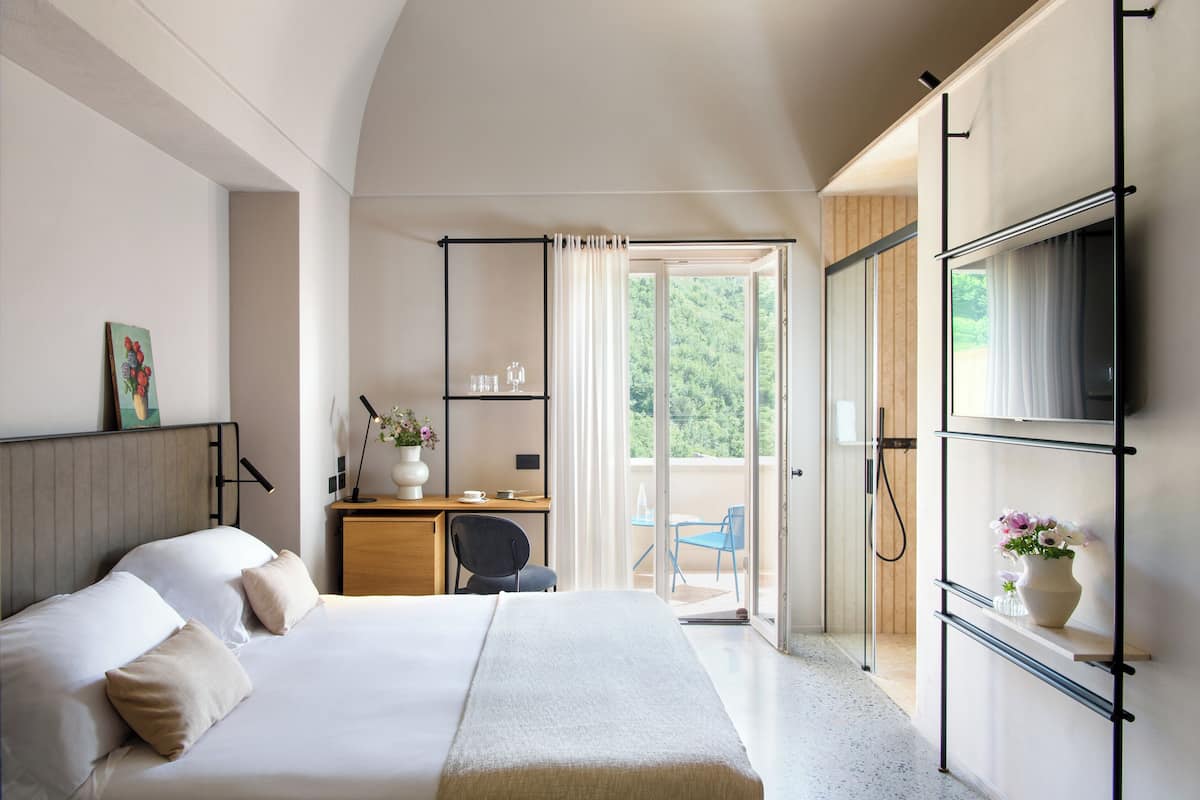 A bedroom with a bed, a desk with a chair, a TV mounted on the wall, and a sliding glass door leading to a balcony.