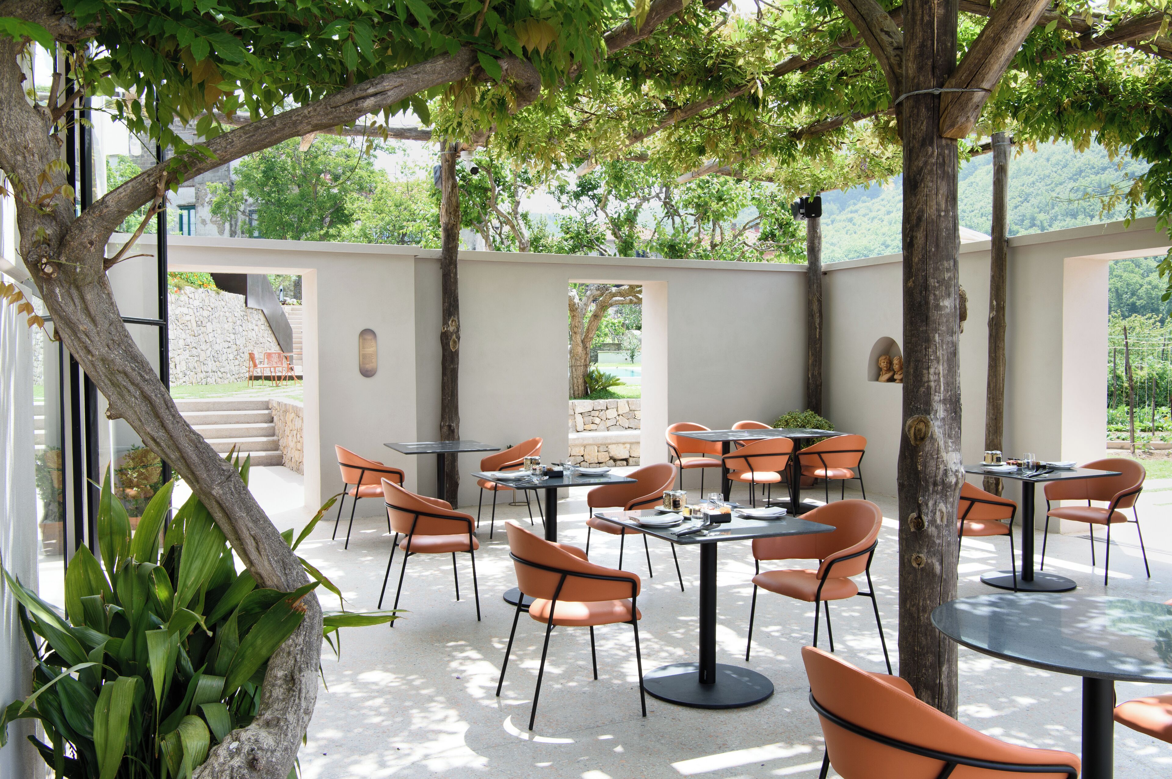 An outdoor dining area with tables and chairs, surrounded by trees and a stone wall.