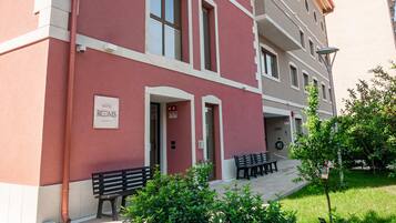 Ein rotes Gebäude mit vielen Fenstern und einem Schild, das es als Hotel kennzeichnet. Vor dem Gebäude befinden sich Bänke und ein kleiner Garten.