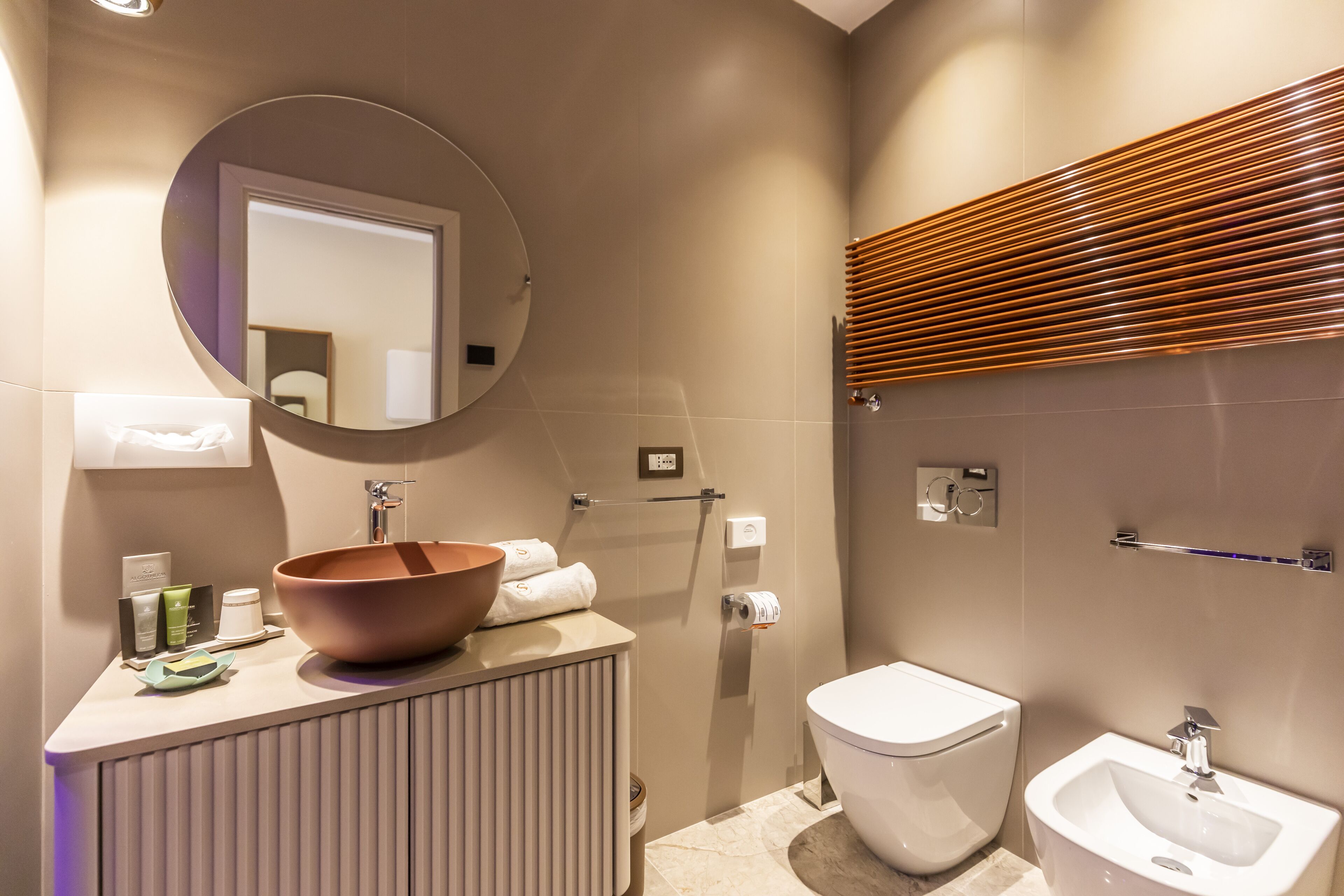 Un baño moderno con un espejo redondo, un lavabo de diseño, un inodoro y un bidé.