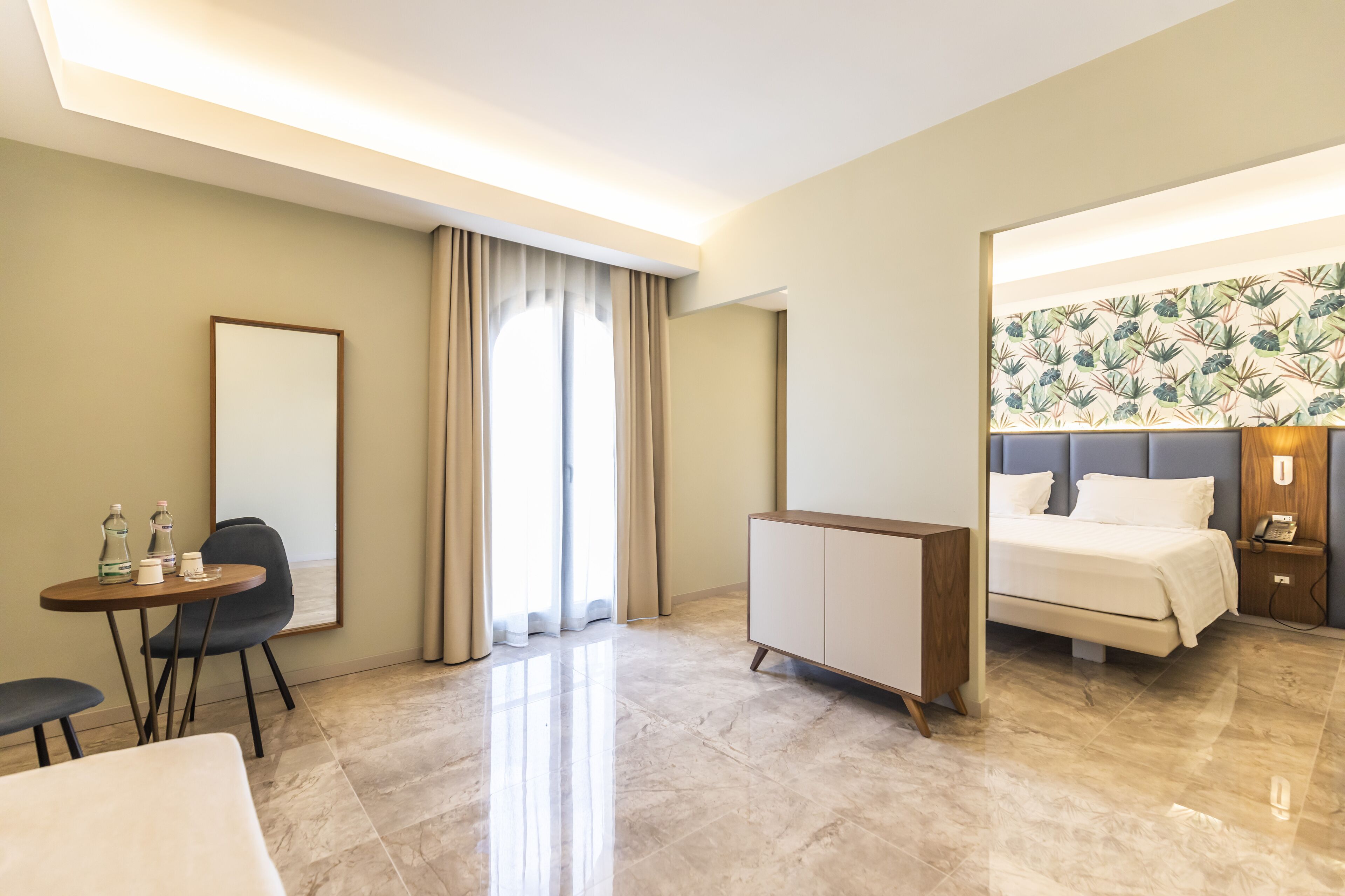A hotel room with a bed, a desk with a chair, a wardrobe, and a large mirror.