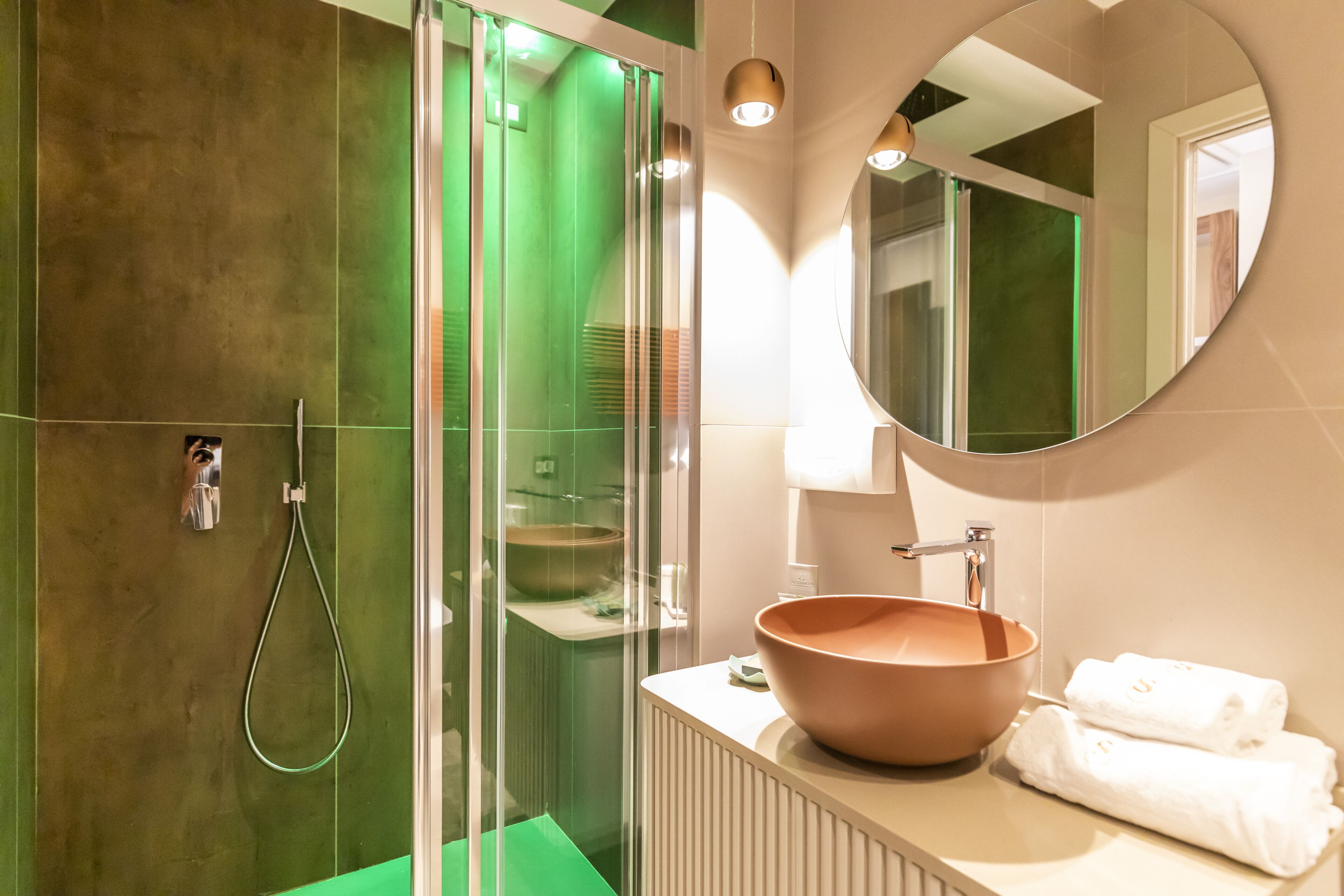 Ein modernes Badezimmer mit einer gläsernen Duschkabine, einem runden Spiegel und einem beigen Waschbecken mit einer großen Schüssel.