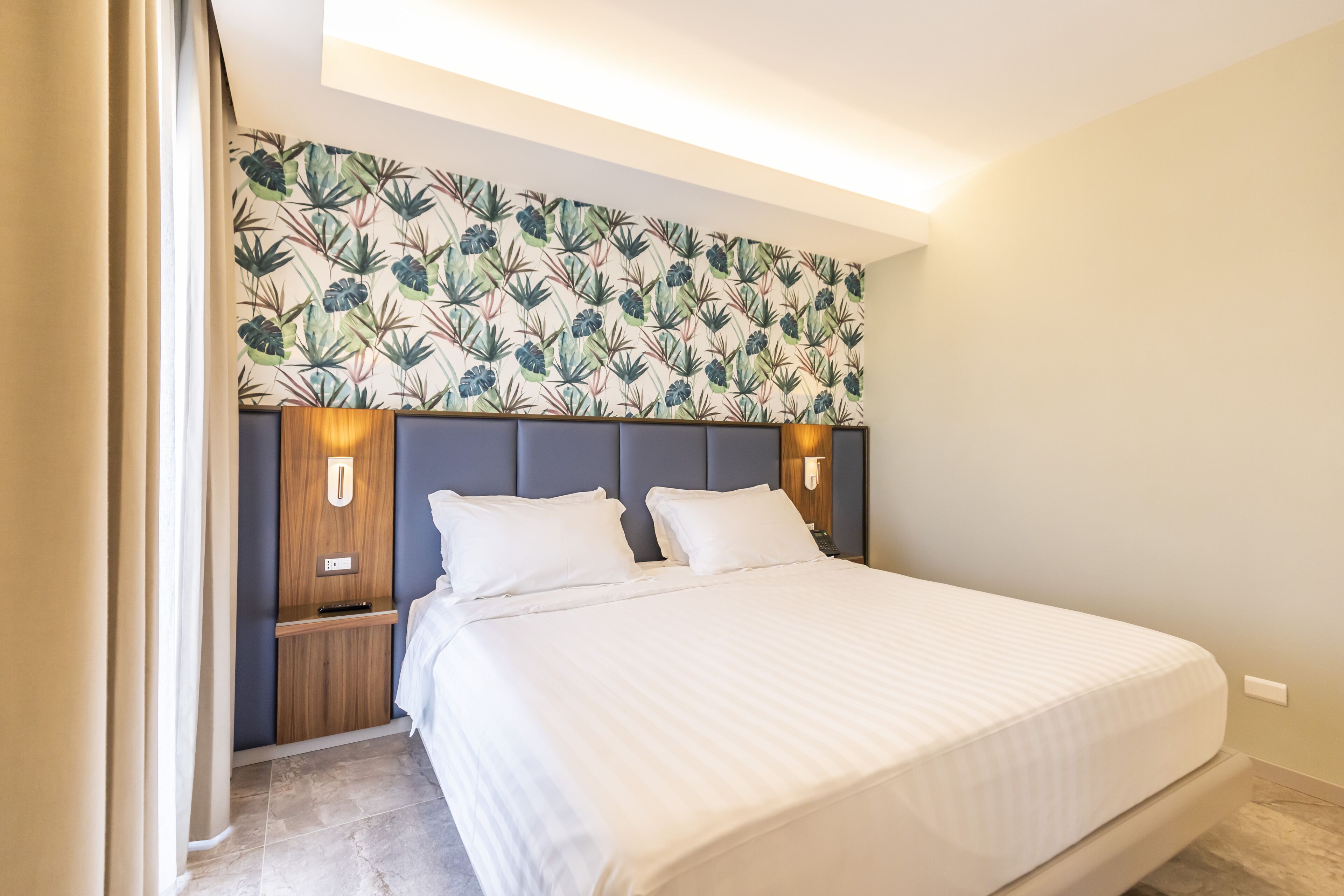 Een hotelkamer met een groot bed, een hoofdbord met behangpapier met een bladmotief, nachtlampjes en een nachtkastje.