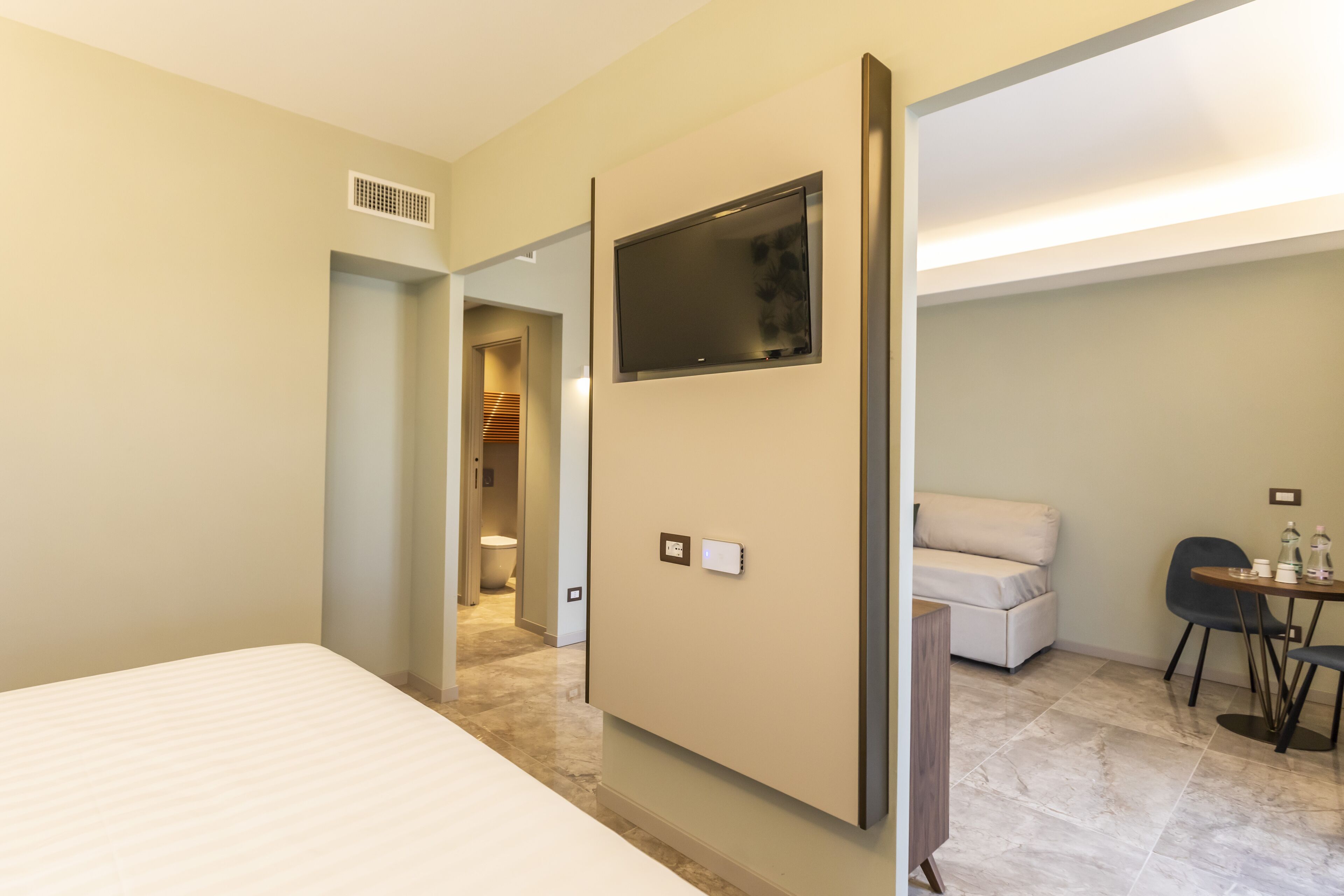 Quarto de hotel com cama, televisão fixada na parede, área de estar com mesa e cadeiras e um pequeno sofá.