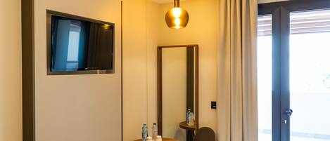 A hotel room with a flat-screen TV mounted on the wall, a round wooden table with two bottles and a cup, a brown chair, a large mirror, and a door with a view of the outside.