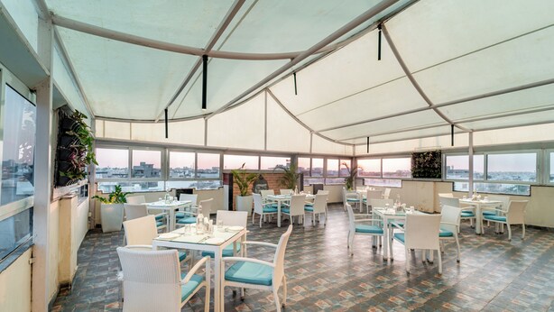 Un amplio espacio de comedor al aire libre con mobiliario de mimbre blanco y cojines azules, ventanas grandes que ofrecen vistas a la ciudad y suelo de baldosas.