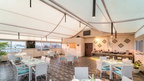 Un amplio comedor interior con mesas y sillas blancas, piso con diseño y un ventanal con vista a la ciudad.