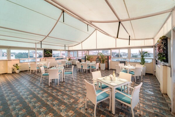 A spacious outdoor dining area with multiple tables and chairs, large windows offering views of the city, and potted plants adding greenery.