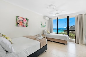 A hotel room with two beds, a ceiling fan, and a large window with a view of the sea.