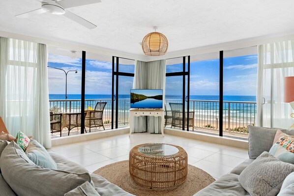 A spacious living room with a large TV, comfortable seating, and a view of the beach through large windows.