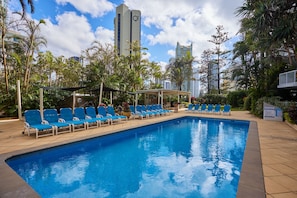 A swimming pool with blue chairs and a covered seating area, surrounded by palm trees and tall buildings.