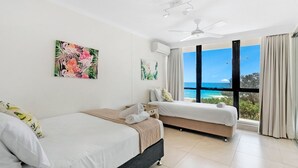 A hotel room with two beds, a ceiling fan, and a large window with a view of the ocean.