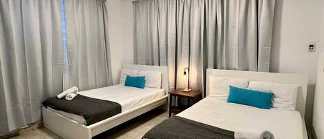Una habitación de hotel con dos camas individuales, una mesita de noche con lámpara y un ventanal con cortinas.