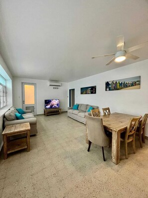 A spacious living room with a sofa, a TV, a dining table with chairs, and a ceiling fan.