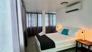 A hotel room with a bed, a bedside table with a lamp, an air conditioning unit, and curtains.