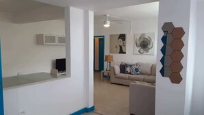 A modern living room with a sofa, a coffee table, and a lamp. There is a kitchen area with cabinets and a countertop.