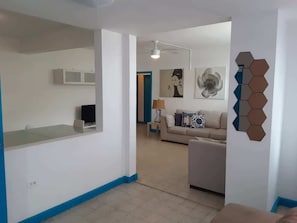 A modern living room with a sofa, a coffee table, and a lamp. There is a kitchen area with cabinets and a countertop.