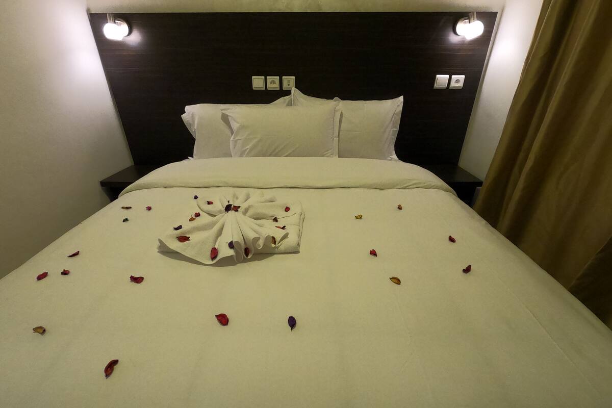 A neatly made bed with a towel arranged in a fan shape, surrounded by scattered rose petals.