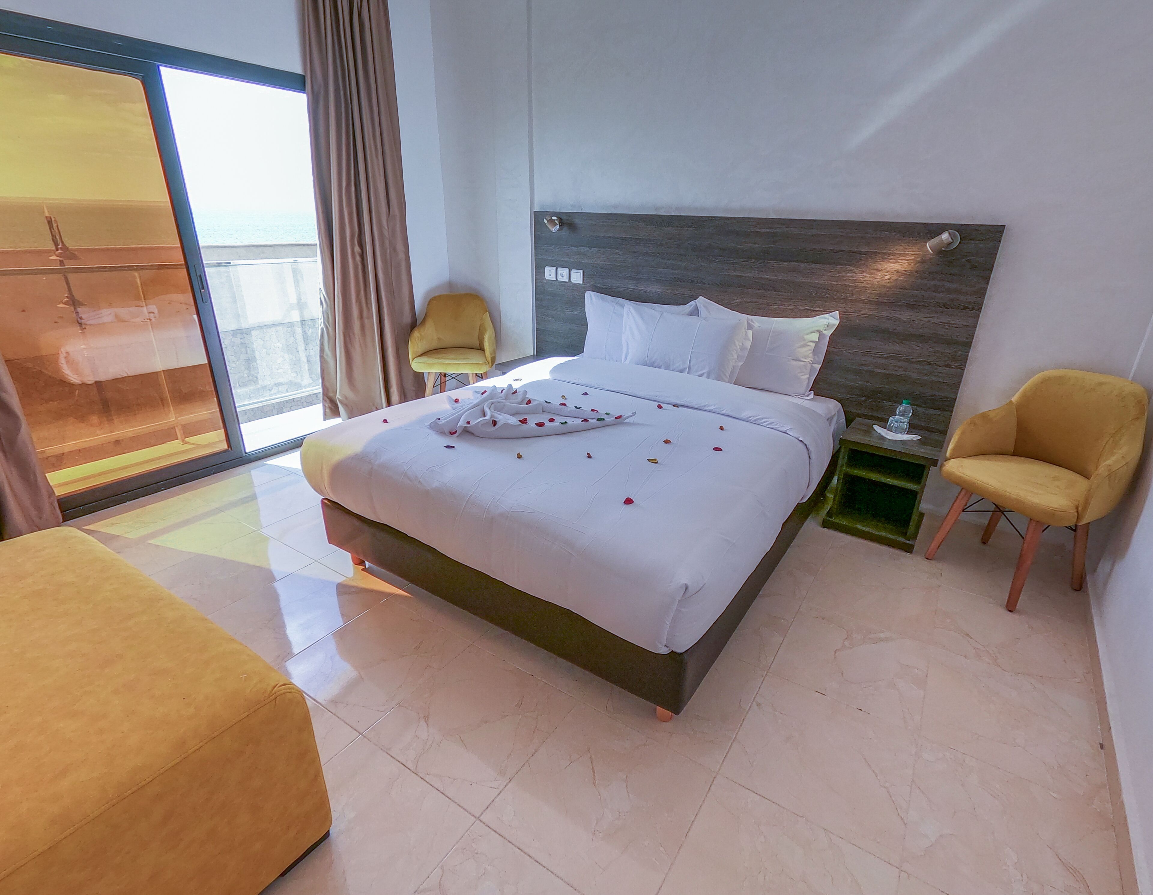 Habitación de hotel moderna con una cama grande, un sillón amarillo, una mesita con una botella y vistas al mar a través de un ventanal amplio.