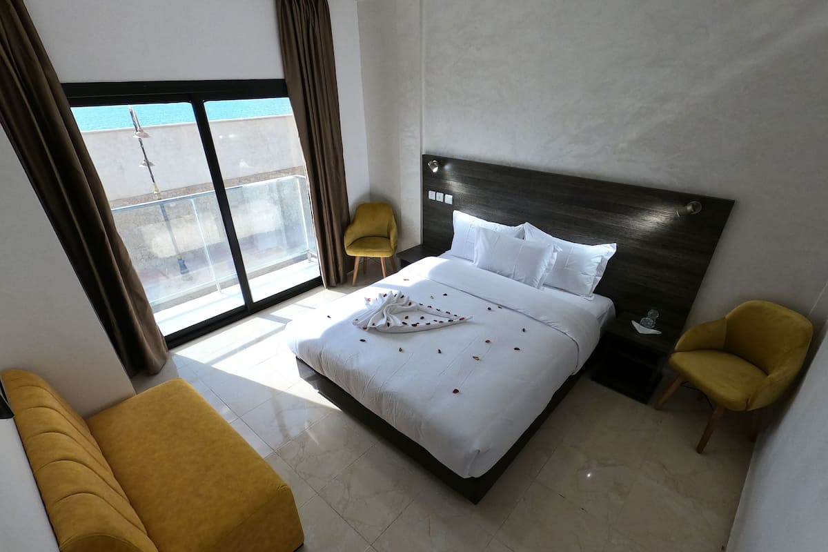 A hotel room with a large bed, a yellow armchair, a small table, and a view of the outside.