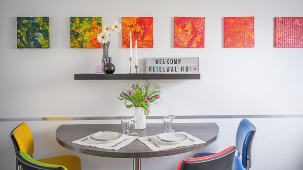 Ein Essbereich mit einem runden Tisch, gedeckt für zwei Personen, einer Vase mit Blumen und einem 'Welcome'-Schild an der Wand.