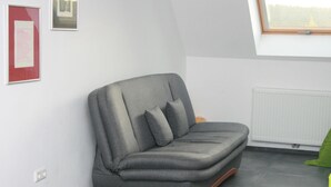 Ein graues Sofa mit geschwungener Rückenlehne und Armlehnen, vor einer weißen Wand mit einem gerahmten Bild links.