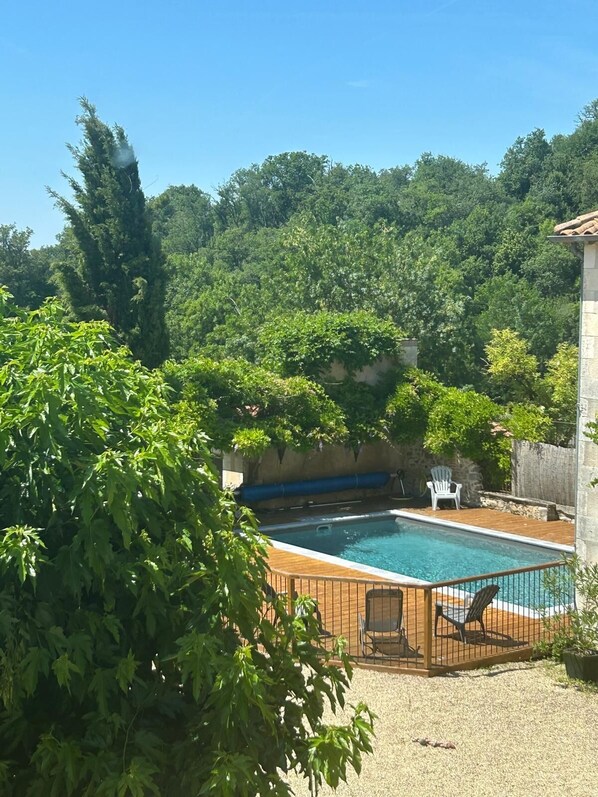A swimming pool with a wooden deck and chairs, surrounded by greenery and a stone wall.