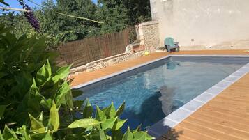 A swimming pool with a wooden deck, surrounded by greenery and a stone wall.