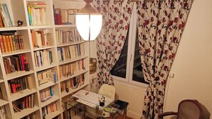Une pièce avec une bibliothèque remplie de livres, un bureau avec une chaise et une fenêtre ornée de rideaux à motifs floraux.