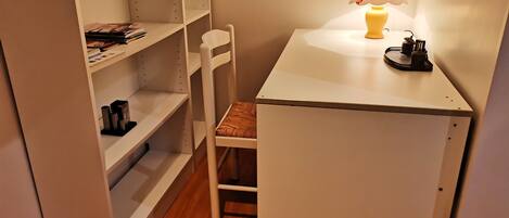 Una habitación pequeña y ordenada con un escritorio blanco, una silla, una estantería y una lámpara.