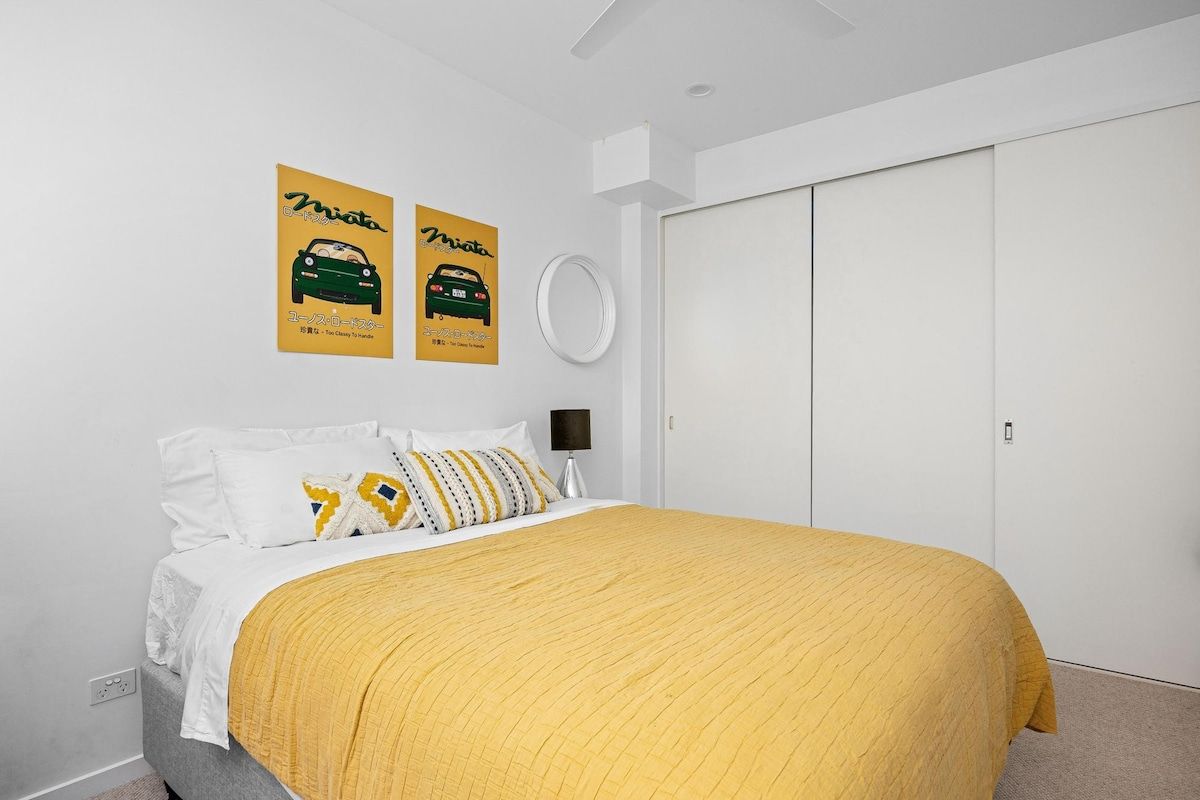 A bedroom with a bed, white walls, a yellow bedspread, and two posters on the wall.