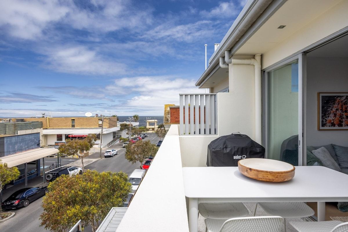 A balcony with a table, chairs, and a grill, overlooking a street with cars and buildings.