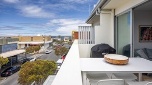 A balcony with a table, chairs, and a grill, overlooking a street with cars and buildings.