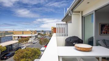A balcony with a table, chairs, and a grill, overlooking a street with cars and buildings.
