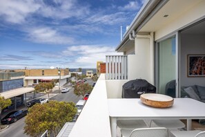 A balcony with a table, chairs, and a grill, overlooking a street with cars and buildings.