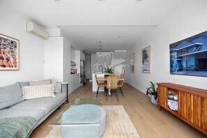 Ein modernes Wohnzimmer mit einer Couch, einem Glastisch, einem Essbereich mit einem Holztisch und Stühlen sowie einer Küche im Hintergrund.
