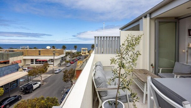 A balcony with a view of the street and ocean, a small potted plant, and a seating area with cushions.