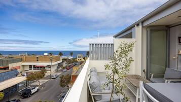 A balcony with a view of the street and ocean, a small potted plant, and a seating area with cushions.
