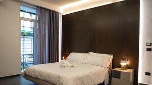 A modern bedroom with a large bed, a nightstand with a lamp, and a view of the outdoors through a window. - Hart Boutique Rooms (Naples)