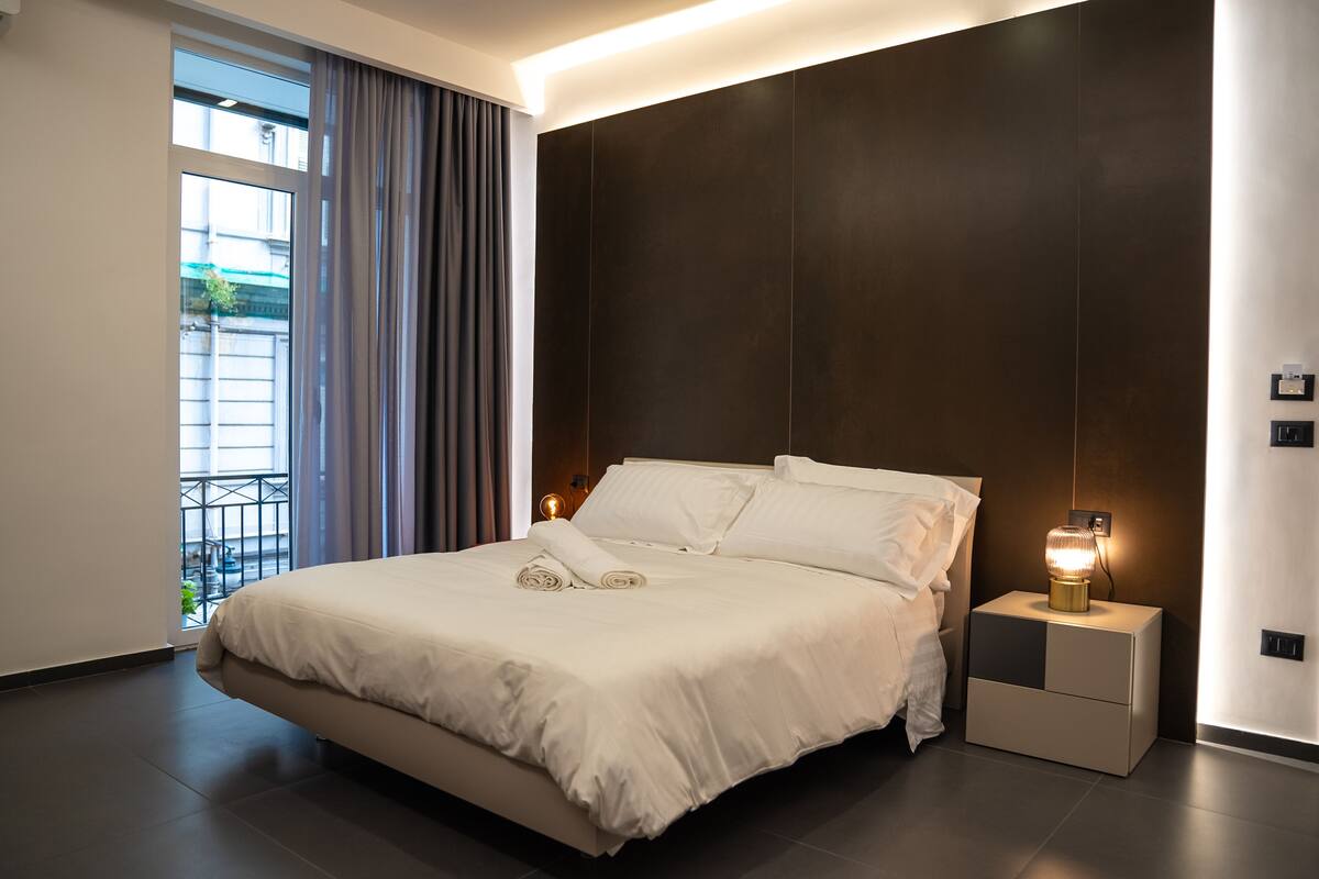 A modern bedroom with a large bed, a nightstand with a lamp, and a view of the outdoors through a window.