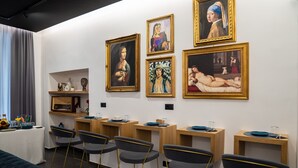 A modern art gallery with paintings on the walls, wooden tables with chairs, and a bed in the foreground. - Hart Boutique Rooms (Naples)