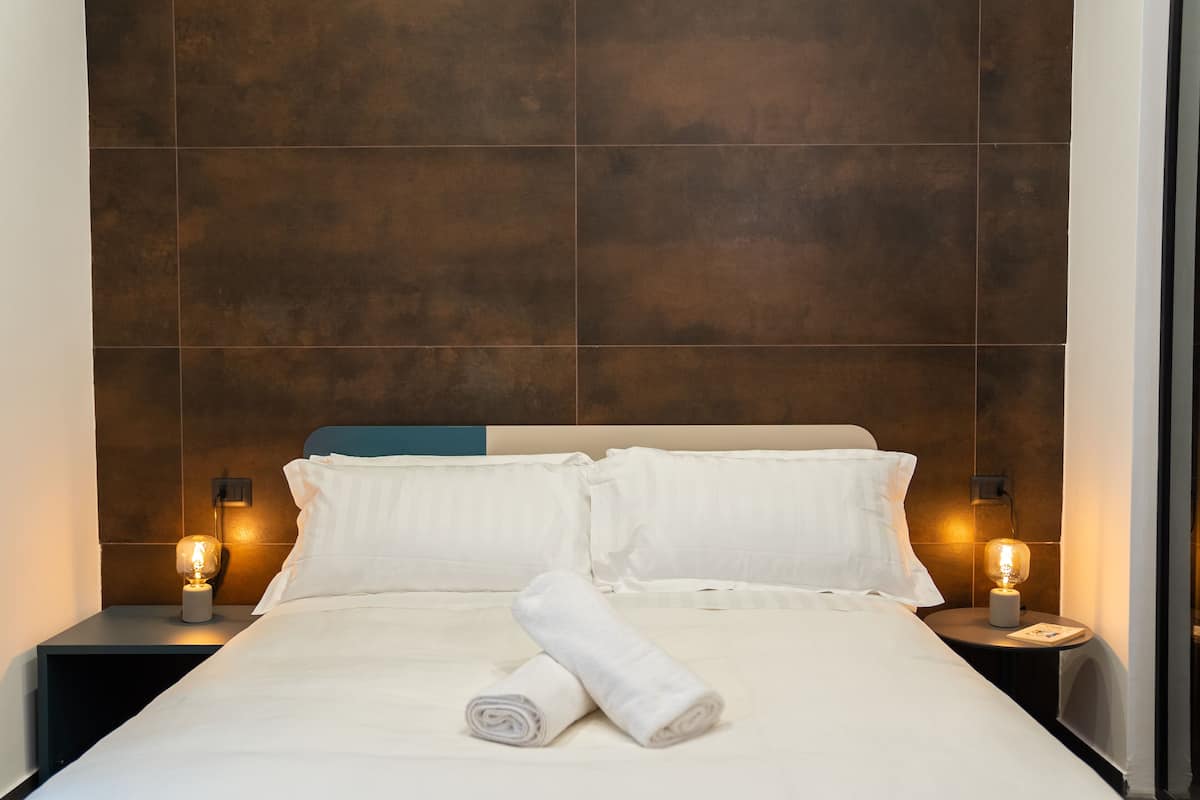 A neatly made bed with white linens and pillows, two bedside lamps, and a rolled towel on the bed.
