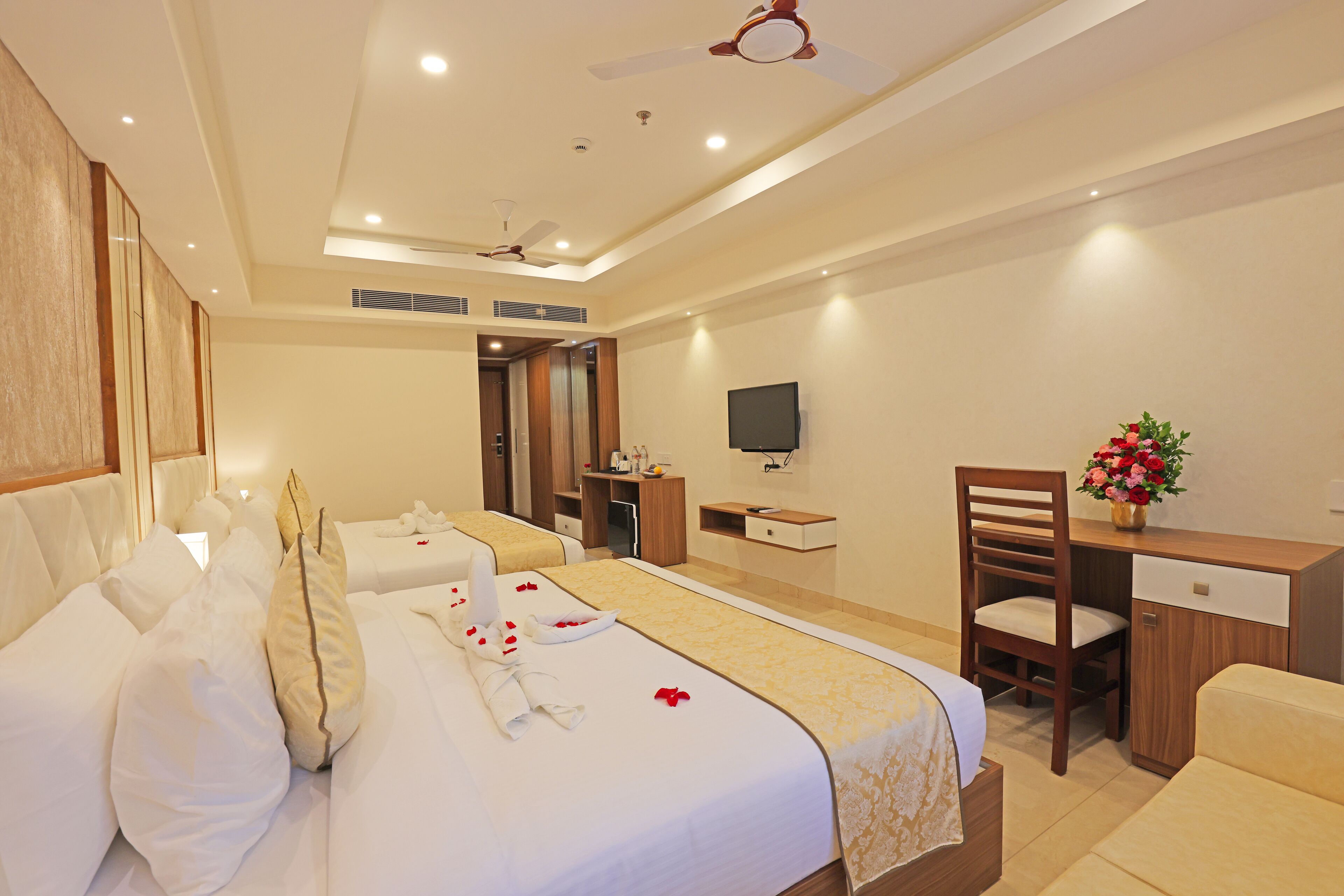 A hotel room with two beds, a desk, a chair, a TV, and a flower vase.