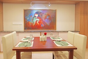 Un salón de conferencias con una mesa de madera dispuesta para cuatro personas, sillas blancas, un cuadro abstracto grande en la pared y una pequeña rosa roja sobre la mesa.