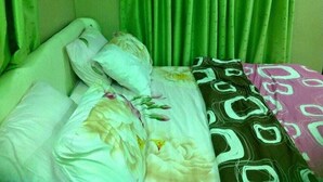 A bed with a patterned blanket and pillows, green curtains, and a plain wall. - Blue-view Suites & Apartments (Abuja)
