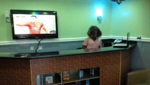 A reception area with a TV mounted on the wall, a counter with a glass top, and a brick-patterned wall. - Blue-view Suites & Apartments (Abuja)