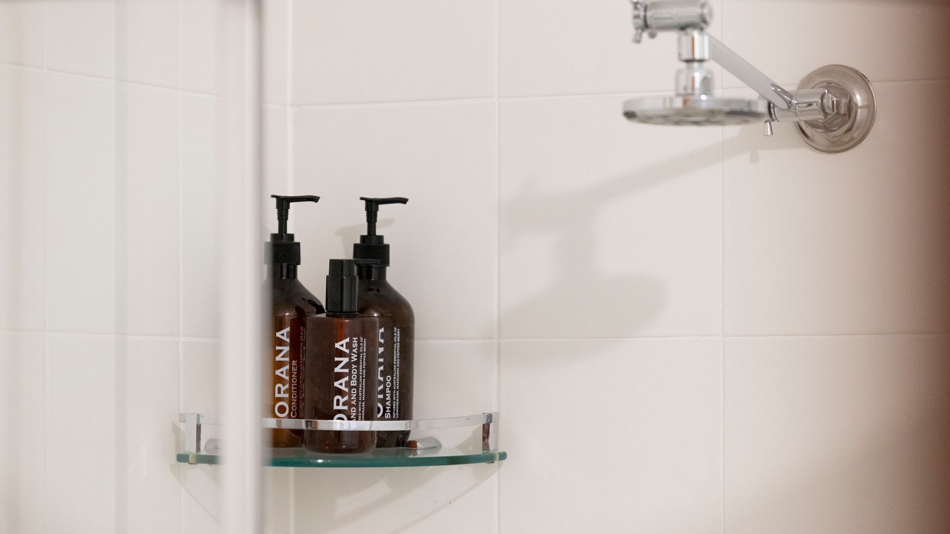 A bathroom with white tiles, a glass shelf holding two bottles of ORANA body wash and shampoo, and a chrome showerhead.
