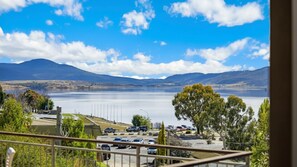 Vista de um lago com montanhas ao fundo, um estacionamento com carros e um edifício com cerca em primeiro plano.