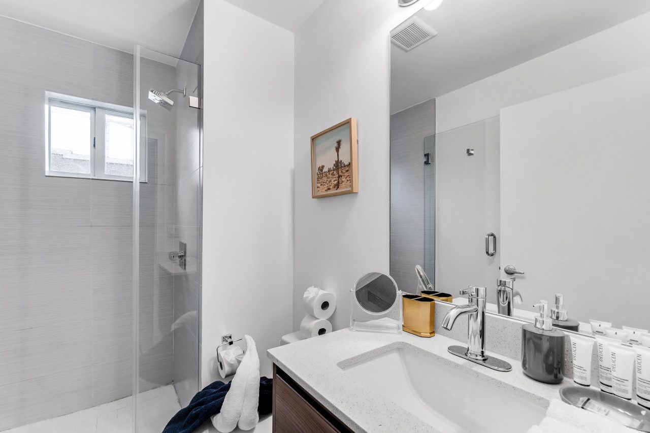 Une salle de bain moderne avec une douche en verre, un grand miroir et un lavabo blanc agrémenté d’articles de toilette.